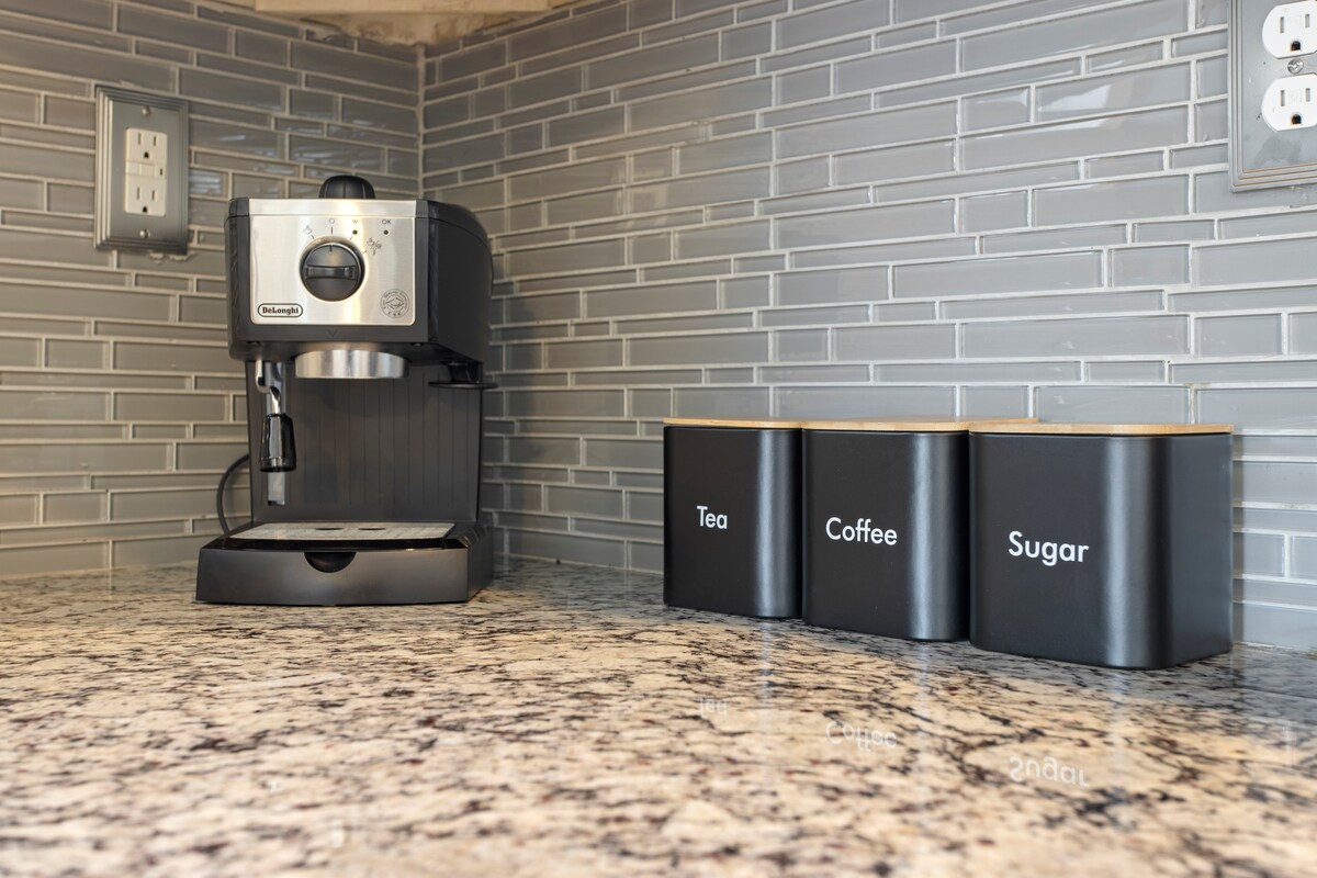 A modern kitchen countertop is showcased, featuring a stainless steel coffee machine alongside three black containers labeled 'Tea', 'Coffee', and 'Sugar'. The countertop is made of speckled granite, complemented by a grey tiled backsplash.