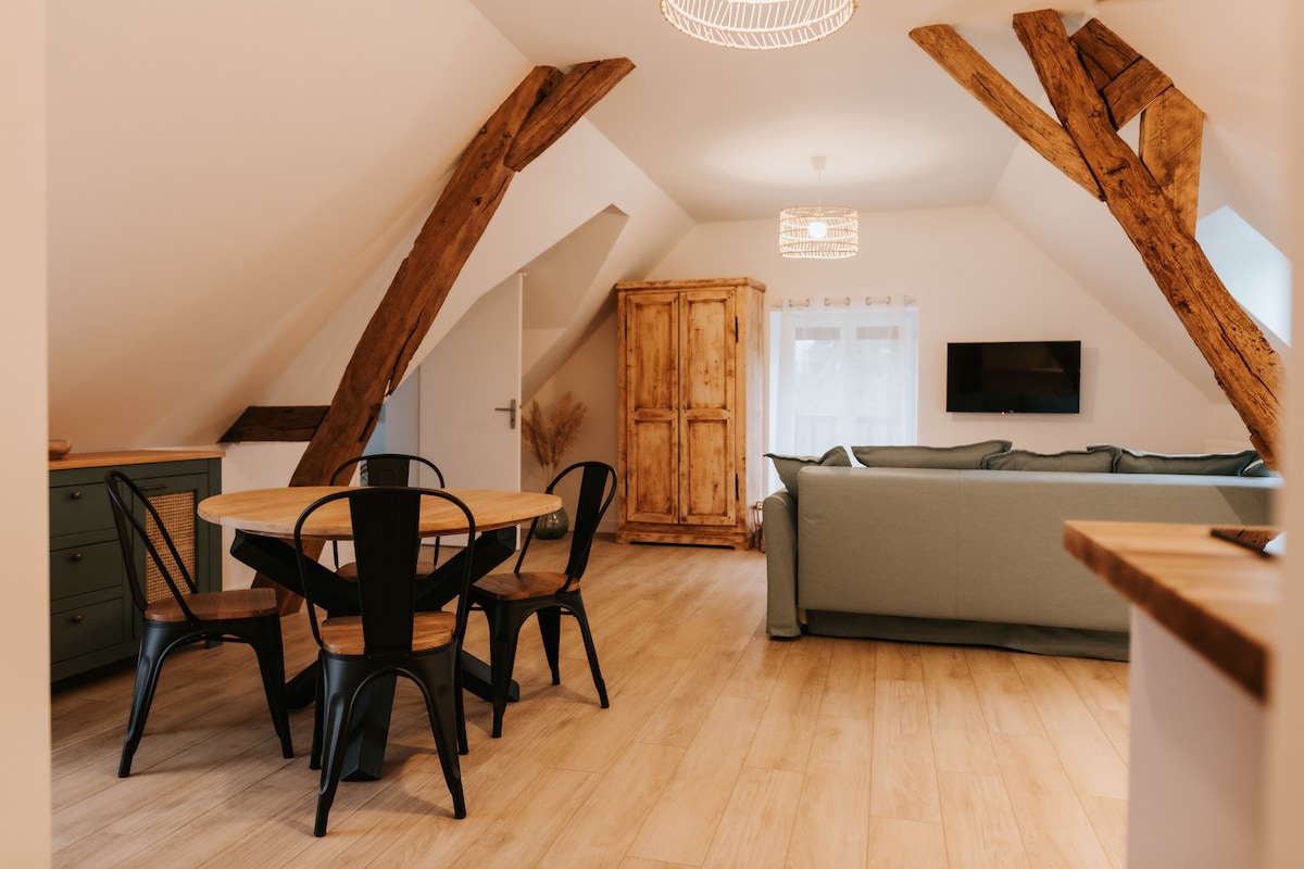 An open living space features exposed wooden beams that add character. A round dining table with black chairs is positioned near a wall, while a light-colored sofa sits opposite a wall-mounted TV. A wooden cabinet adds a rustic touch to the area.