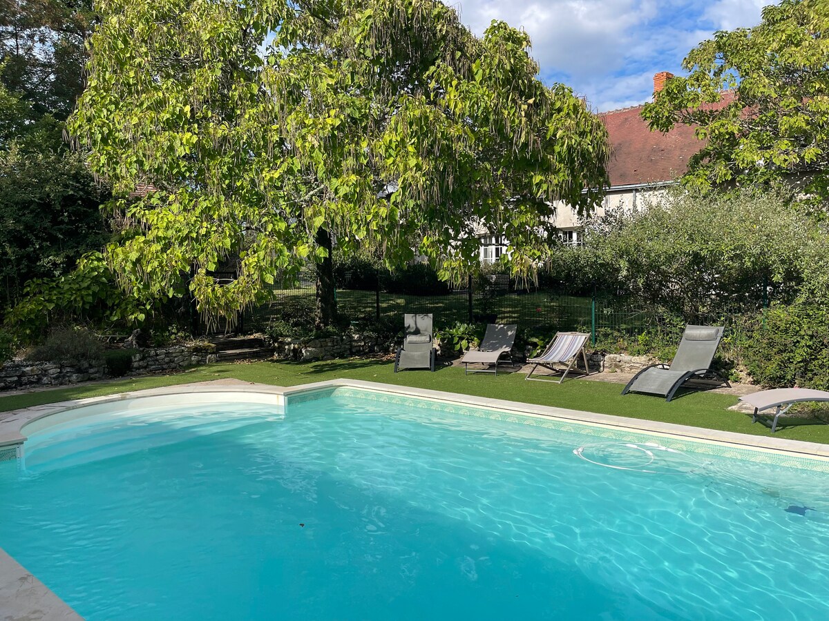 A peaceful outdoor area features a refreshing swimming pool surrounded by lush greenery. Sun loungers are placed nearby, providing a comfortable spot for relaxation. A serene garden setting enhances the inviting atmosphere, with a view of the accommodation's exterior in the background.