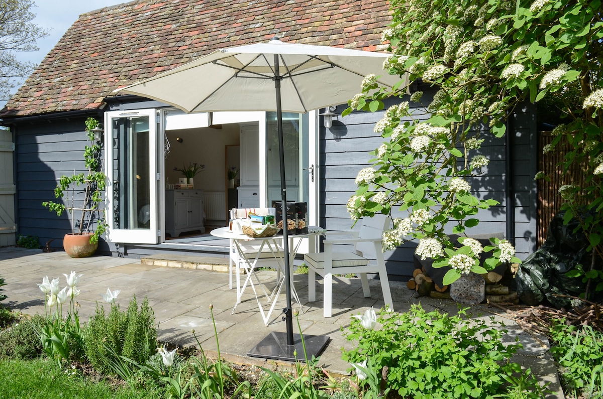 A patio area features a round table and white chairs, shaded by a large umbrella. The entrance to the barn is visible through open glass doors, revealing a glimpse of the interior. Lush greenery and flowering plants frame the scene, enhancing the tranquil outdoor setting.