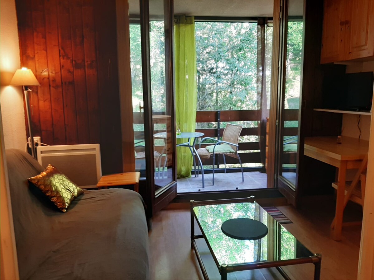 A bright living area features a sofa bed and a glass coffee table. Large windows open to a loggia furnished with a small table and chairs, offering views of the surrounding trees and nature.