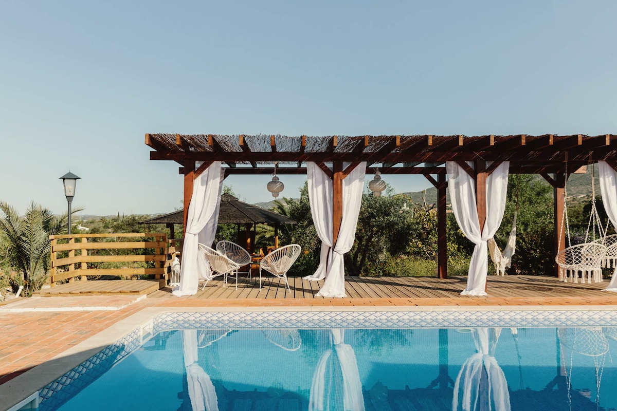A shaded terrace features draped white curtains, gently billowing in the breeze, alongside two wicker chairs. The inviting salt-water pool reflects the clear blue sky, surrounded by lush greenery that enhances the tranquil outdoor setting.