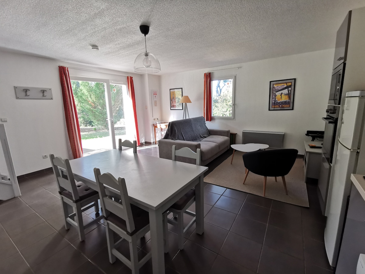 A spacious living area presents a dining table with six white chairs, complemented by a gray sofa. Large windows with light curtains overlook a verdant outdoor space. A modern coffee table and an array of kitchen appliances, including a refrigerator and an oven, are visible.