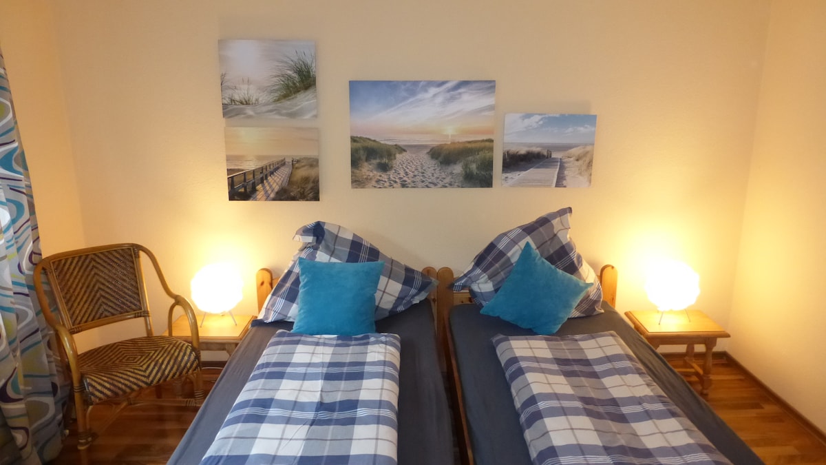 The sleeping area features two separate beds adorned with blue and gray checkered bedding. Each bed is complemented by plush turquoise pillows. Flanking the beds are two nightstands with soft glowing lamps. Above the beds, three landscape images depict serene beach scenes.