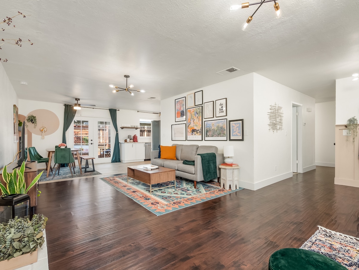 An open living area features a spacious layout with hardwood flooring and a decorative area rug. Comfortable couches in muted tones are arranged around a central coffee table, while wall art and plants add character. Access to natural light is provided through sliding glass doors leading to the patio.