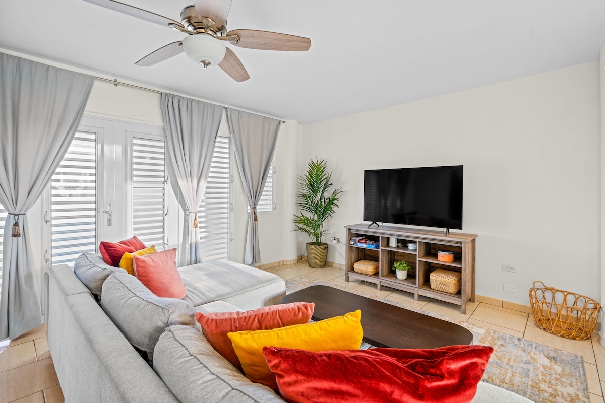 A light-filled living space features a spacious sectional sofa adorned with colorful cushions. A large flat-screen TV is positioned on a rustic wooden media console. Light gray curtains frame the windows, while a potted plant adds a touch of greenery.