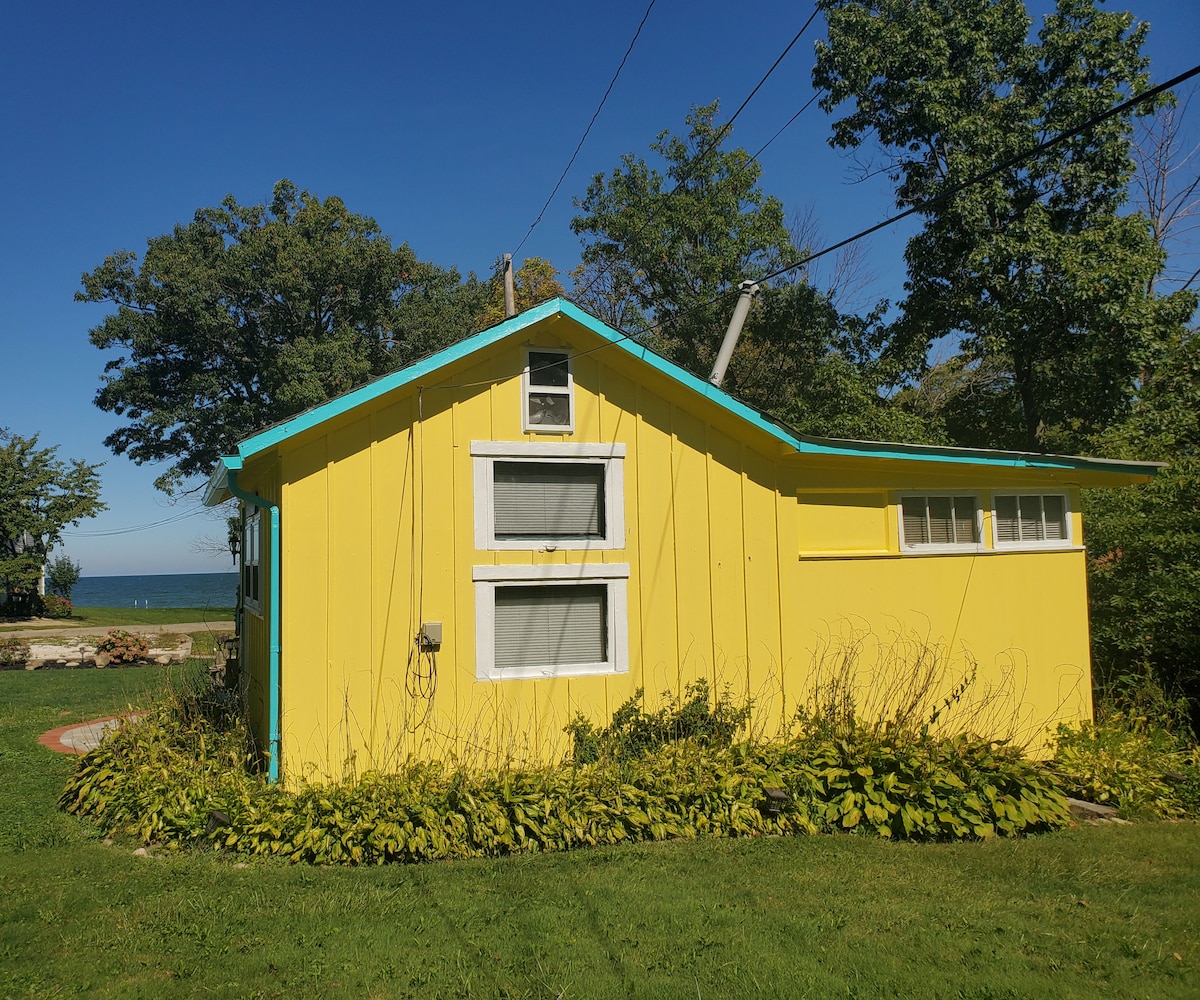 The exterior of a bright yellow cottage is shown, featuring turquoise accents. Large windows provide glimpses of the surroundings. Lush greenery and flowering plants surround the base, while the expansive Lake Erie is visible in the background under a clear blue sky.