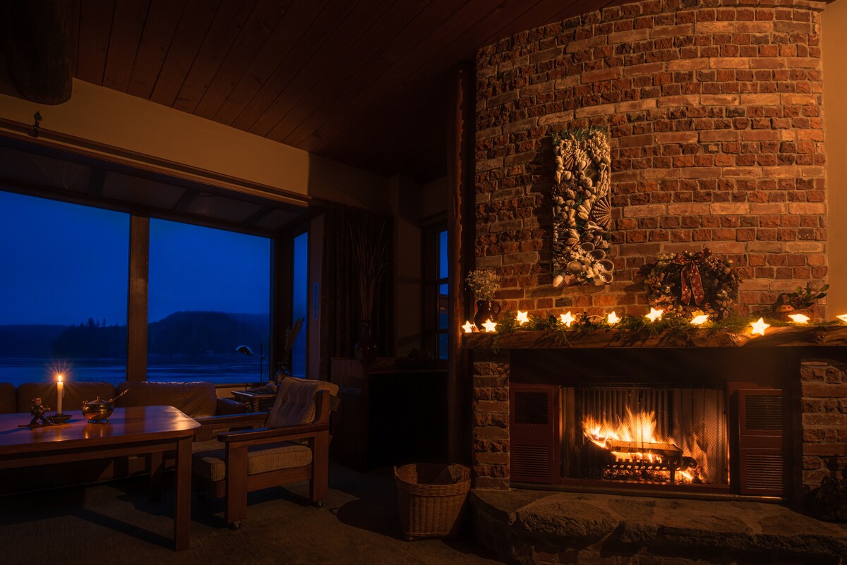 A cozy living area is highlighted by a brick fireplace with a warm fire. Soft lighting from candles decorates the mantel, and large windows offer views of the evening landscape. Comfortable seating is arranged nearby, inviting relaxation in the tranquil setting.