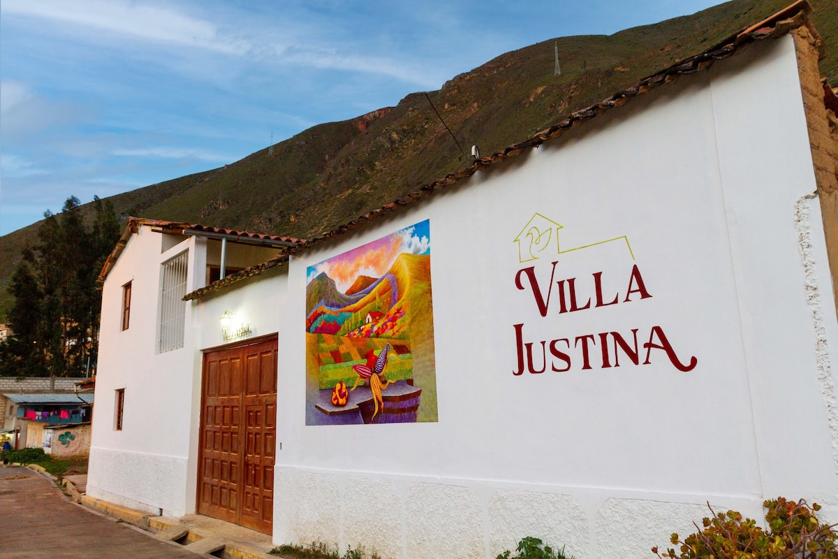 The exterior of Villa Justina features a white facade with a colorful mural depicting local scenery. Wooden double doors provide entry, while the surrounding landscape includes rolling hills in the background. The rustic charm of the building reflects its natural materials and cultural significance.