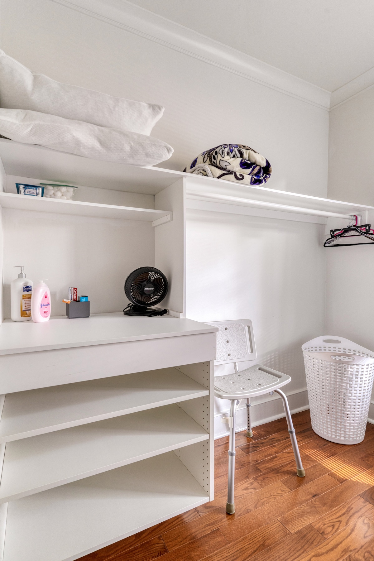 A small closet space features built-in white shelves and a hanging rod. A white chair is positioned beside a small fan on a countertop. Personal items are neatly arranged, including toiletries and a laundry basket. Natural light enhances the warm wooden floor.