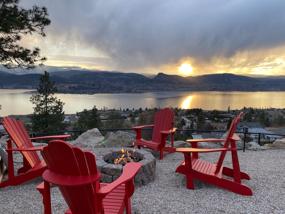A cozy fire pit area features several red Adirondack chairs arranged in a circle. The setting overlooks a tranquil lake reflecting the sunset, surrounded by rolling hills and a peaceful sky. Gravel pathways lead to the seating area, enhancing the natural landscape.