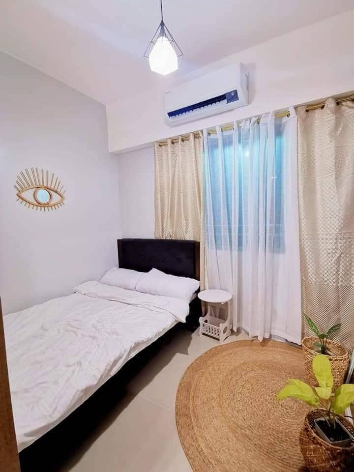 A cozy bedroom is featured with a black bedframe and white bedding. A round jute rug is positioned on the floor. Curtains in soft beige filter natural light from the window, which is adorned with a decorative element on the wall and a small plant in a basket.