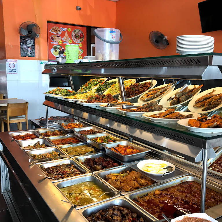 A well-arranged buffet counter displays a variety of dishes in stainless steel trays. Colorful cooked meats and vegetables are presented alongside rice and sauces. Behind the counter, a wall features menus and a list of culinary options for guests to choose from.