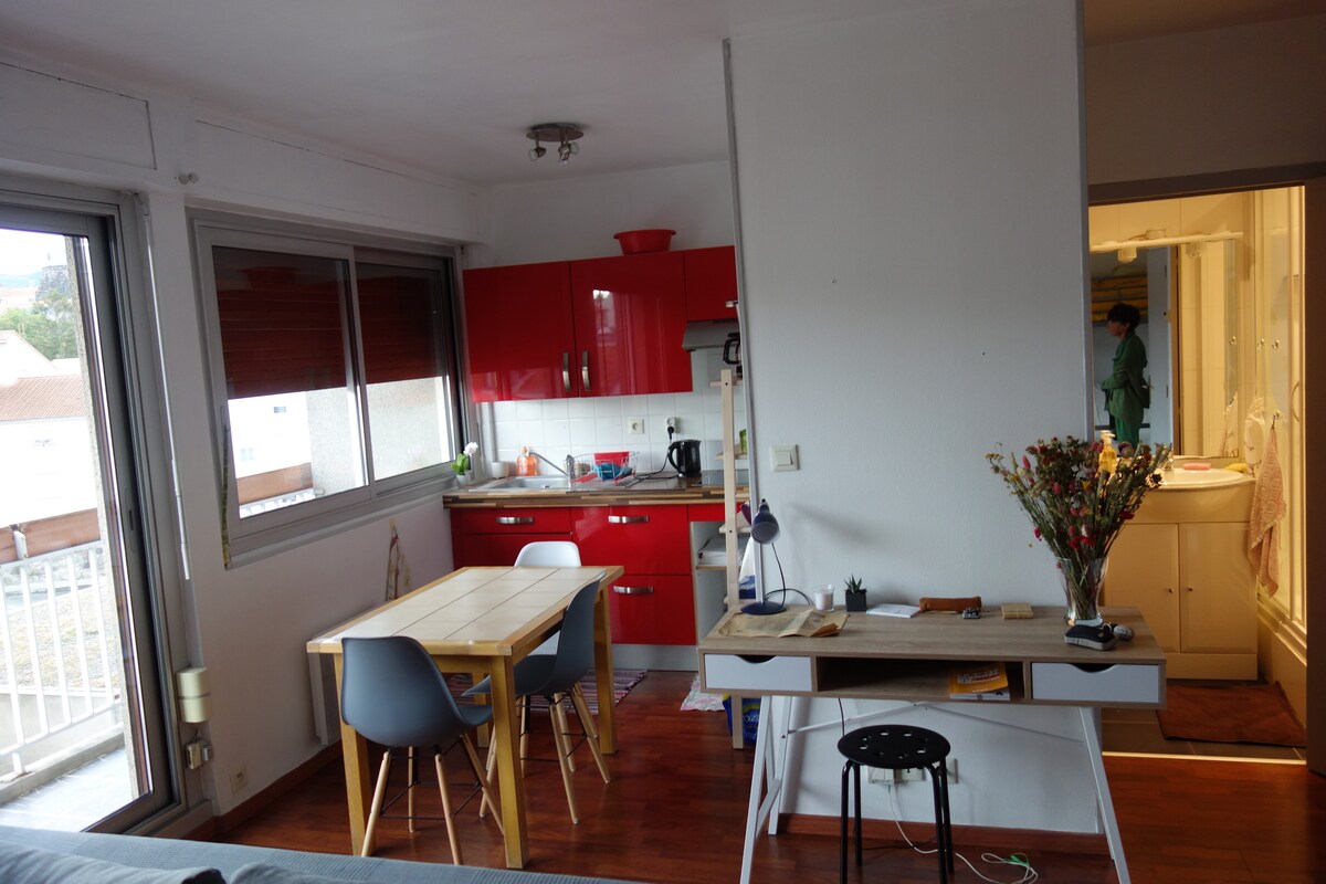An open-plan studio features a compact kitchenette with red cabinetry and modern appliances. A wooden dining table and four chairs provide a casual dining space. A desk with a stool is situated near a window, allowing for natural light. Light wood flooring complements the bright atmosphere.