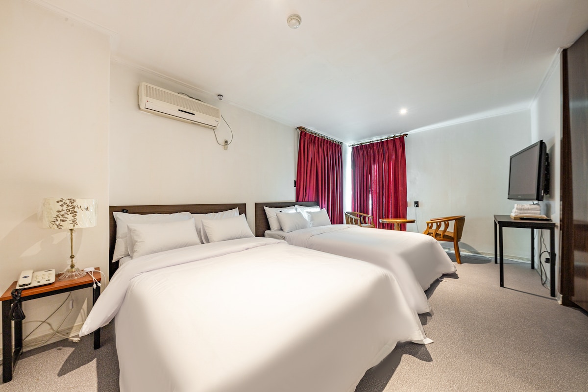 The room features two spacious beds dressed in light linens. Natural light enters through large windows with red curtains. A seating area includes two wooden chairs, and a flat-screen television is positioned against the wall. Air conditioning is available for guest comfort.