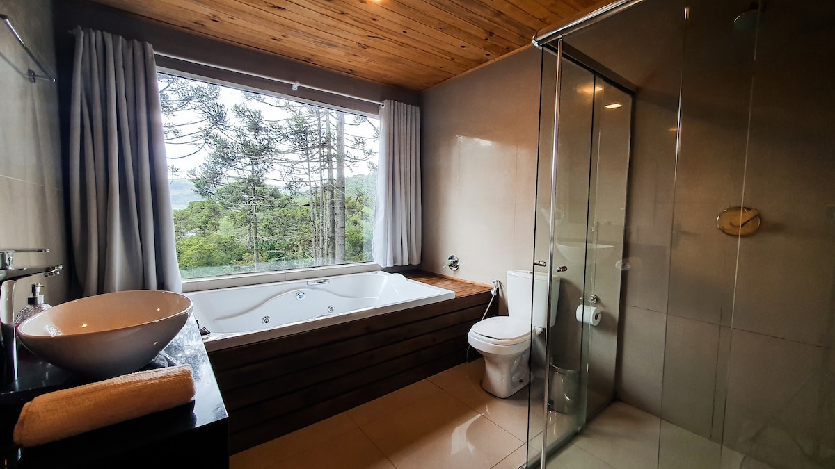 A modern bathroom is showcased with large windows offering views of the surrounding greenery. A spacious whirlpool bath is positioned alongside natural wood accents, while a glass shower stall provides additional functionality. A sleek vessel sink and contemporary fixtures are complemented by soft towels nearby.