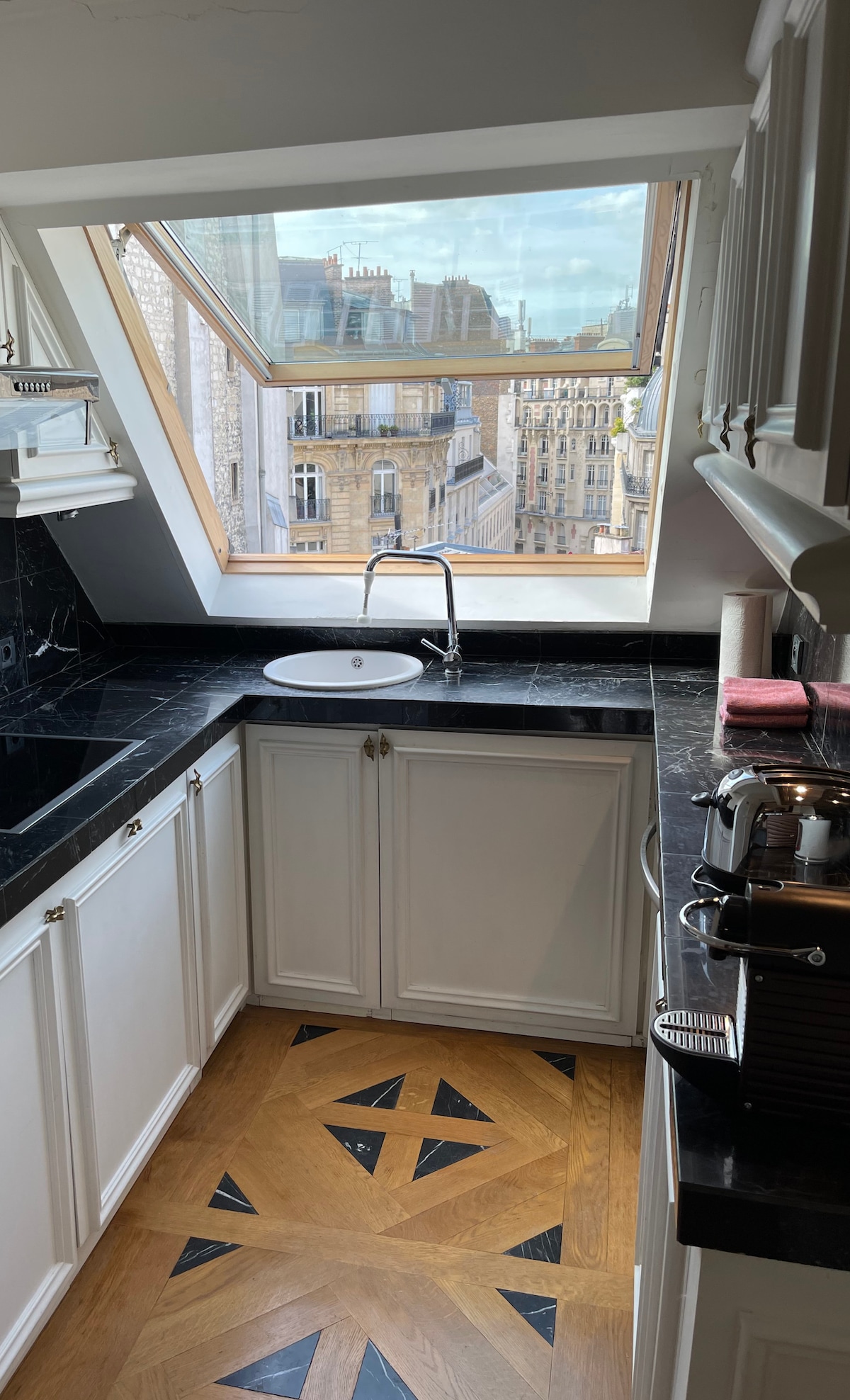 A modern kitchen is showcased, featuring a sleek black countertop and white cabinetry. A unique patterned floor adds character to the space. A triangular window allows natural light to fill the room, providing a view of the city skyline.