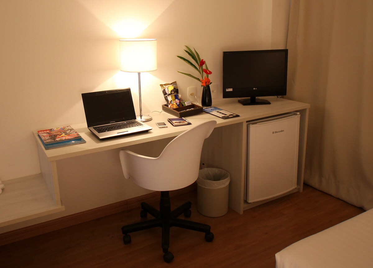 A work station is set up with a laptop on a light-colored desk. A small TV is positioned nearby, alongside a mini-fridge. A decorative plant and a lamp provide subtle lighting, while a comfortable chair completes the workspace.