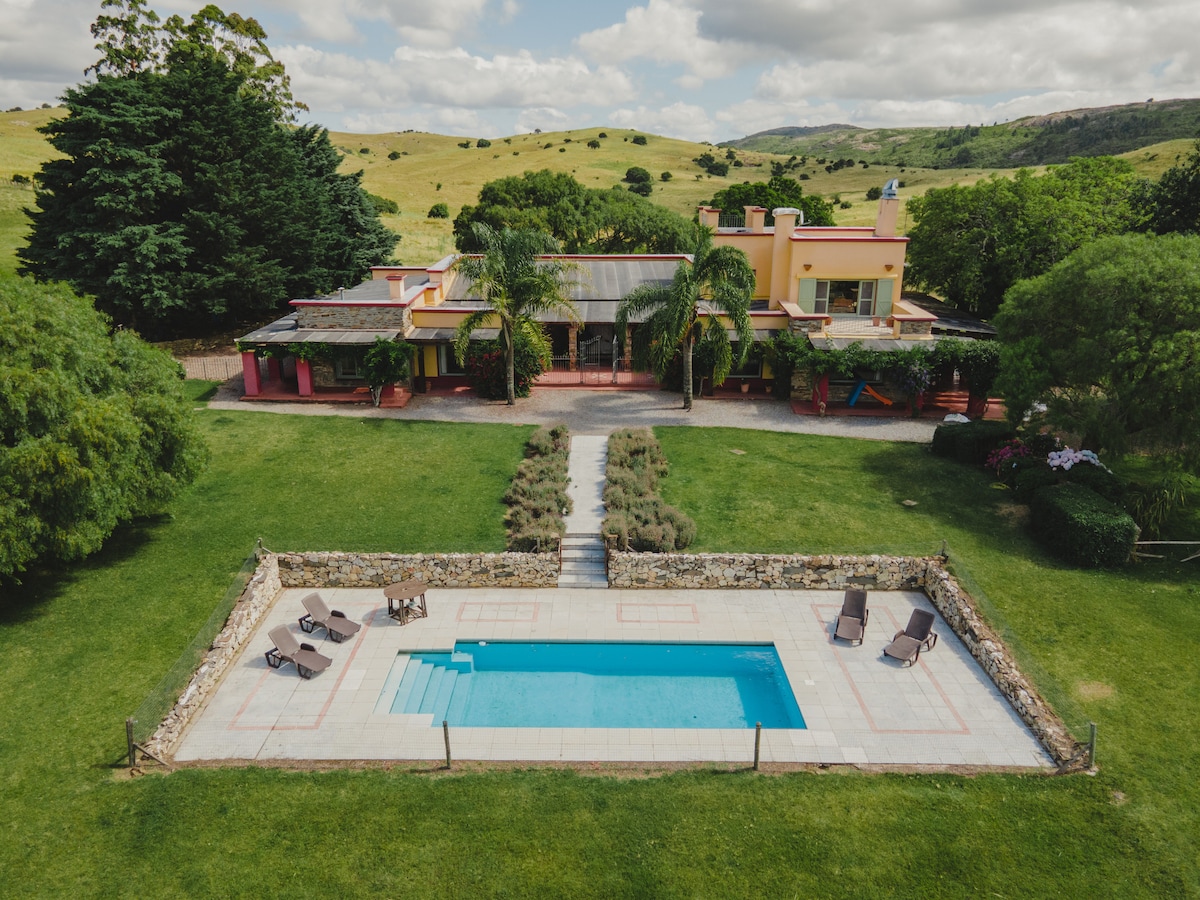 The exterior view captures a spacious house surrounded by lush greenery, set against rolling hills. A crystal-clear swimming pool is centrally positioned, flanked by several lounge chairs. Well-maintained pathways lead through the manicured lawn, inviting relaxation and outdoor activities.