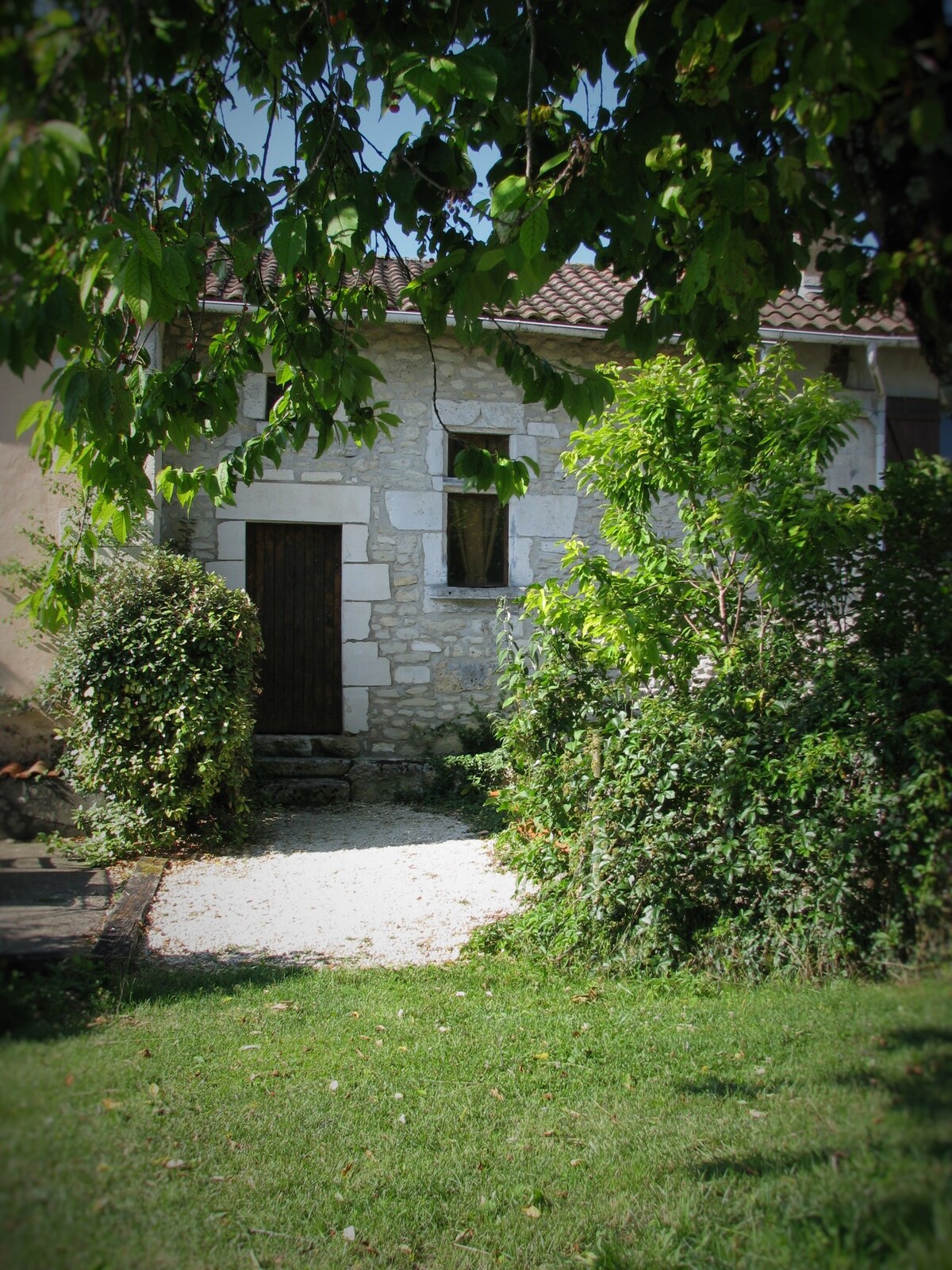 The entrance of a rustic stone gite is framed by lush greenery, including shrubs and trees. A pathway leads to a wooden door, surrounded by a natural landscape that enhances the property's peaceful country setting.