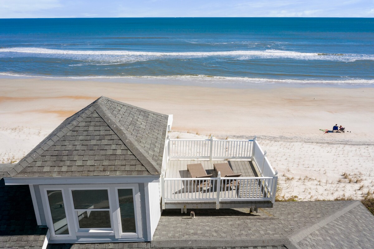 Aerial view showcases a beachfront property with expansive ocean views. A spacious deck with outdoor seating overlooks the sandy beach, where gentle waves can be seen lapping at the shore. The vibrant blue ocean stretches into the horizon, providing a serene backdrop.