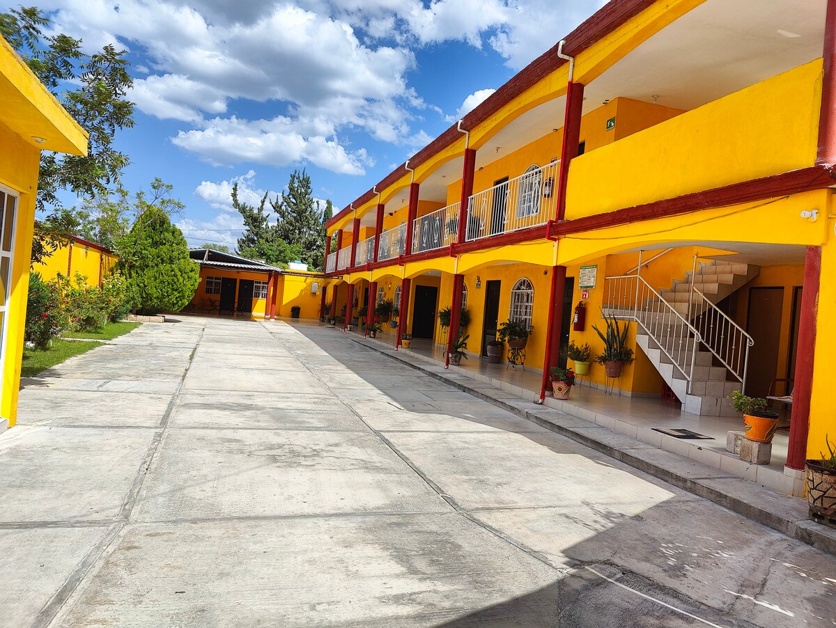 A vibrant yellow building is surrounded by green landscaping. A spacious driveway runs through the center, connecting multiple entrances. Flower pots with plants are placed along the walkway, and a staircase leads to an upper level, enhancing accessibility.