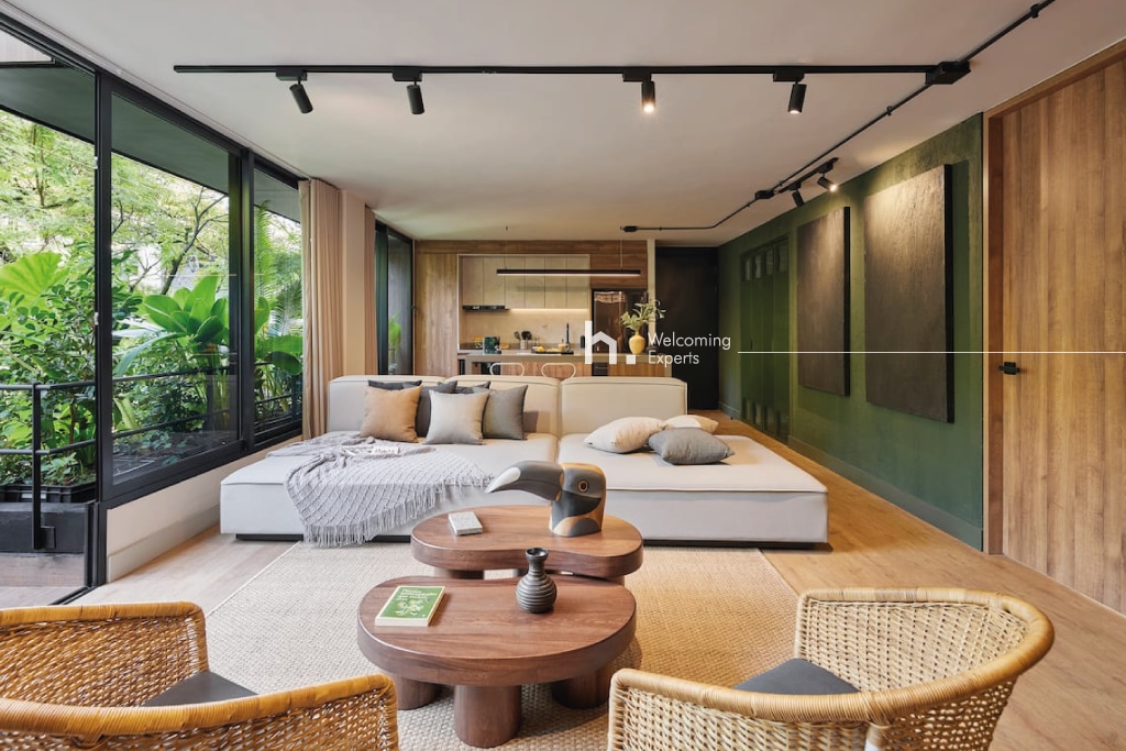 A spacious living area is furnished with a large sectional sofa and two rattan chairs. A wooden coffee table sits at the center, complemented by decorative items. Natural light streams through expansive glass windows, providing views of lush greenery outside. Elegant design features are visible throughout.