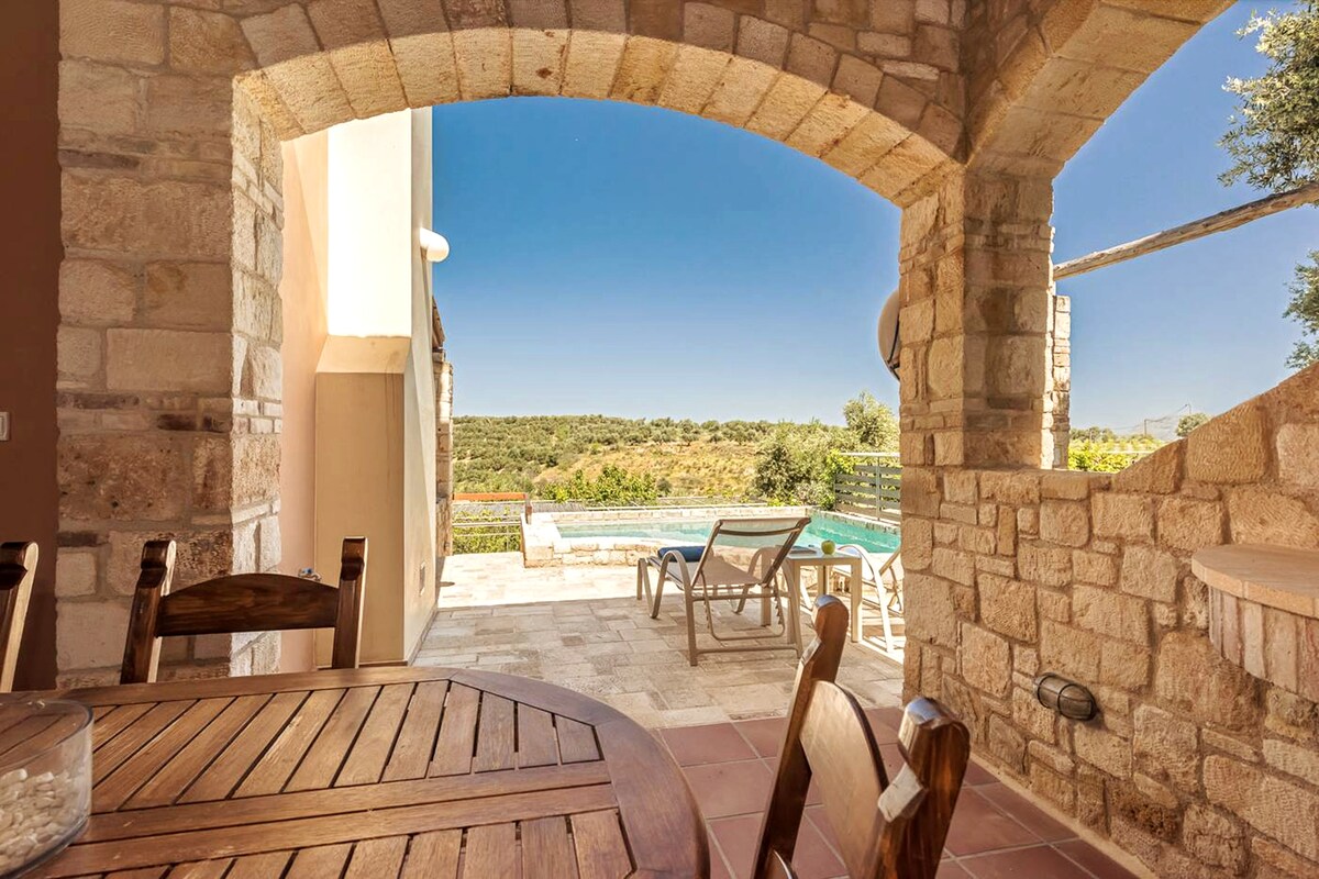 The outdoor area features a spacious stone terrace framed by an archway. In the foreground, a wooden dining table is visible, offering seating for guests. Beyond, a private swimming pool glistens under the sun, surrounded by lounge chairs and olive groves in the background.