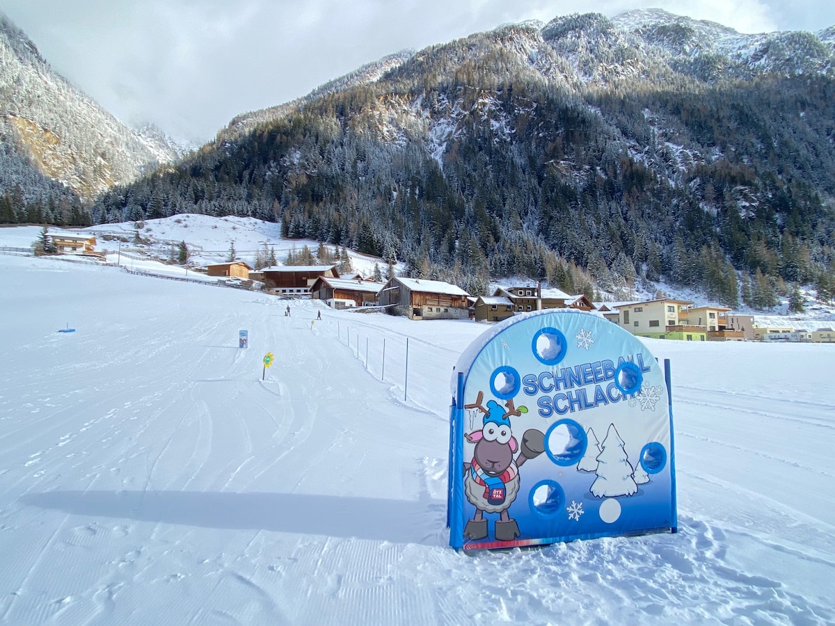 A snow-covered landscape is displayed, featuring a vibrant blue sign with a cartoon character. Surrounding mountains are visible in the background, alongside a collection of traditional chalets nestled in the snow. The scene exudes a serene winter atmosphere.