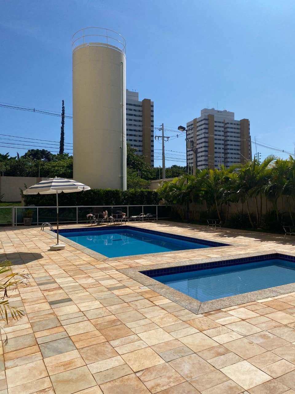A spacious outdoor area features two swimming pools, surrounded by tiled decking. Sun loungers and an umbrella are placed nearby. Lush greenery lines the perimeter, with modern buildings visible in the background under a clear blue sky.