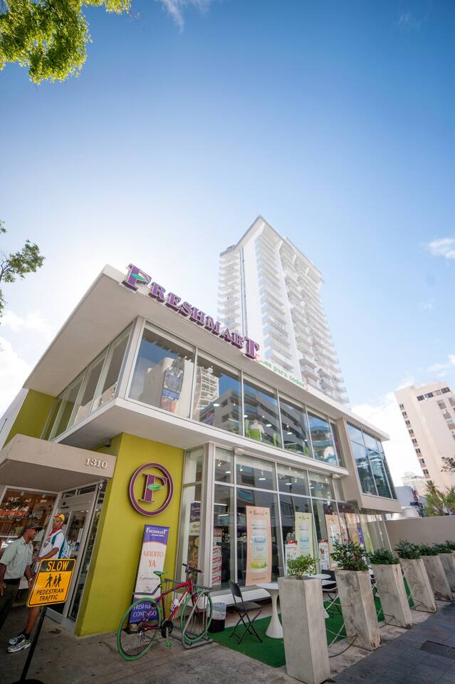 A modern building with a vibrant green exterior features large glass windows. The FreshMart logo is prominently displayed above the entrance. A bicycle is parked outside, and potted plants line the pathway, contributing to the lively atmosphere of the bustling street below.