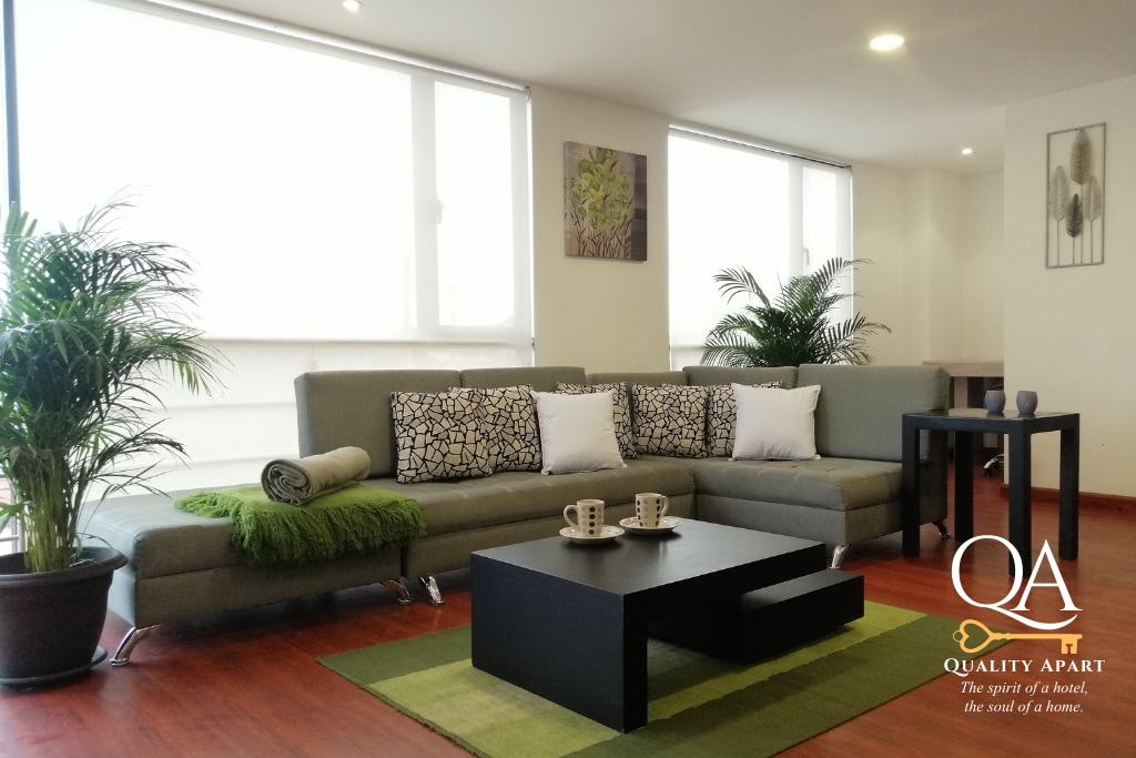 A bright and spacious living room features a comfortable sectional sofa adorned with patterned cushions. A low coffee table is positioned on a green area rug, accompanied by two cups and a small plant. Natural light floods the space through large windows.