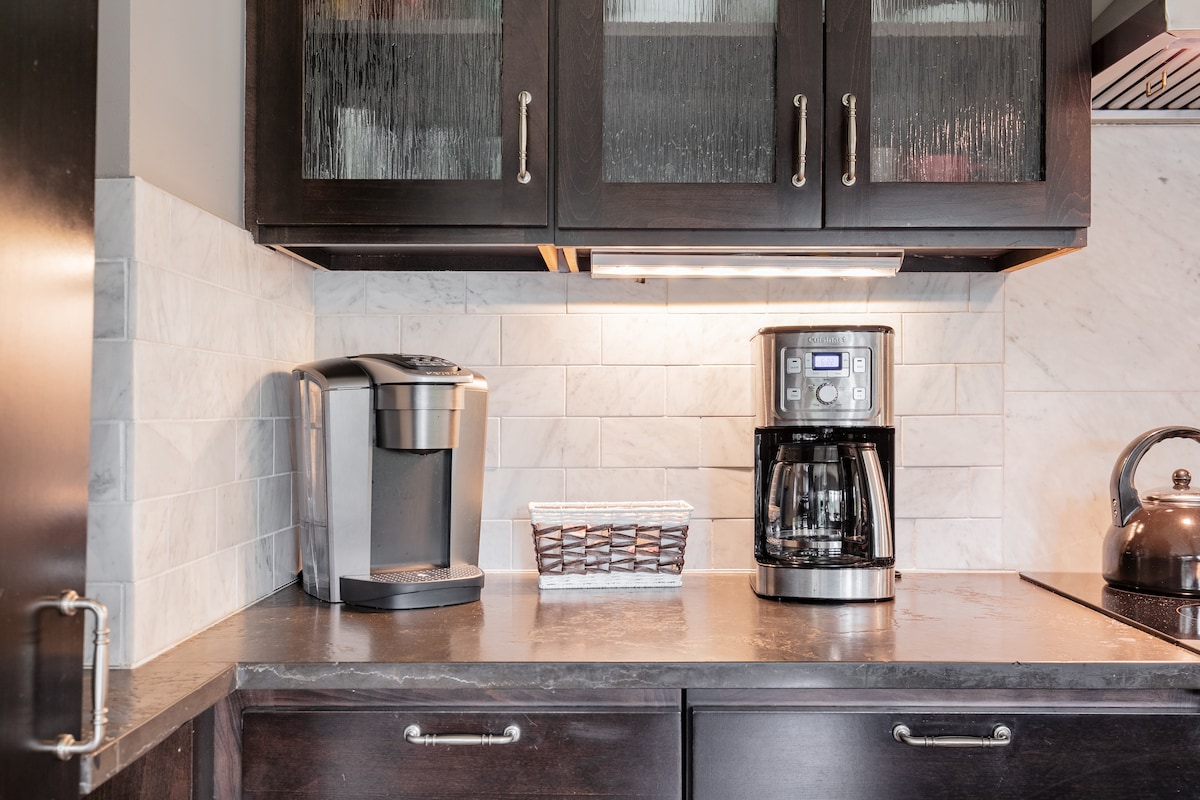 A modern kitchen countertop features two coffee makers, including a single-serve machine and a traditional drip coffee maker. A woven basket is placed nearby, and cabinetry above showcases frosted glass doors, contributing to the kitchen's sleek design.