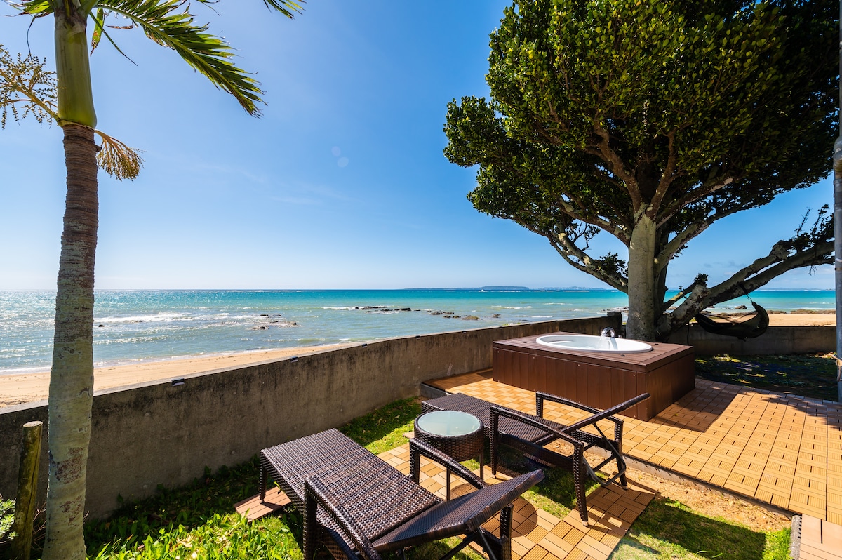 A spacious outdoor area features a hot tub surrounded by a wooden deck. Two rattan chairs and a small table provide a relaxing spot. Lush greenery and a palm tree enhance the setting, with a clear view of the sandy beach and blue ocean in the background.