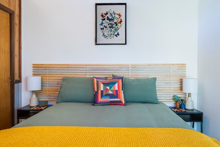 Kitanda cha Malkia katika chumba cha kulala cha ghorofani #2 kinaangalia mwonekano wa miti kupitia mlango mkubwa wa kioo unaoongoza kwenye sitaha ya upande wa kujitegemea. 