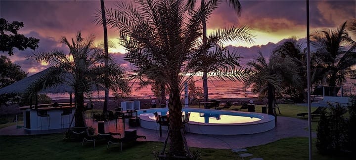 A serene outdoor space is shown featuring a swimming pool surrounded by palm trees. The sunset casts vibrant colors in the sky, reflecting on the water's surface. Lounge chairs are arranged nearby, and distant ocean waves are visible, creating a tranquil beachside atmosphere.