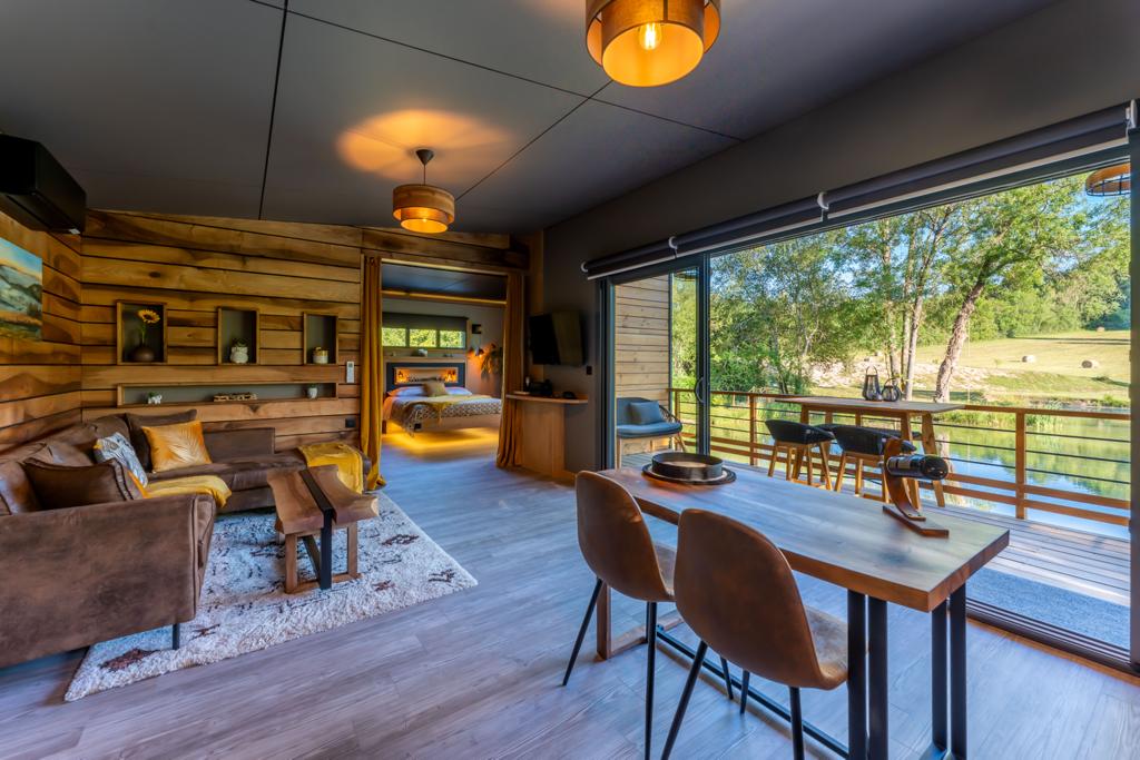 An open living space includes a cozy seating area with a large brown sofa and a wooden coffee table. A dining table with two chairs is visible next to glass doors that lead to a balcony, offering views of the surrounding nature.