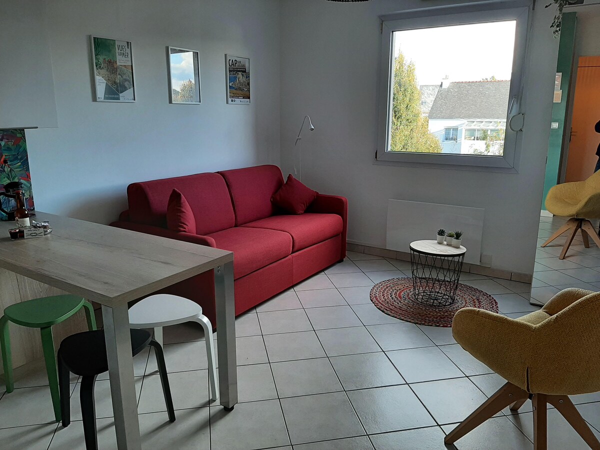 A bright living area features a red sofa positioned near a large window that offers ample natural light. A round coffee table is displayed on a textured area rug, alongside two accent chairs in yellow and green, and a wooden dining table with black and white seating.
