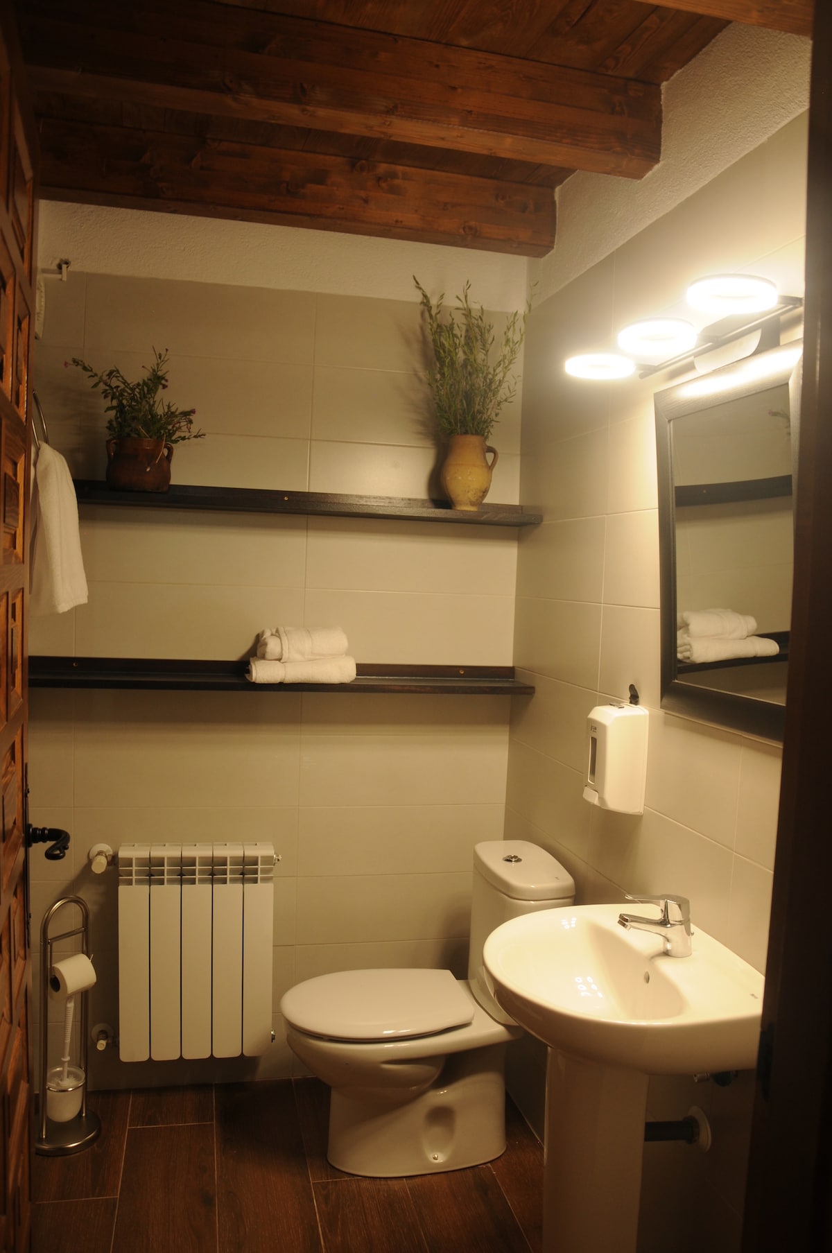 The bathroom is equipped with a white toilet, a modern sink, and neatly arranged toiletries. Floating shelves display fresh towels and decorative plants. A mirror above the sink is illuminated by soft overhead lighting. Warm wooden beams accent the ceiling, adding a touch of texture.