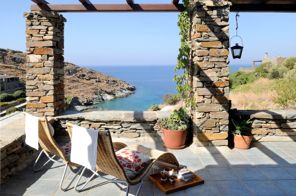 A serene outdoor space features two lounge chairs with patterned cushions, positioned to overlook the coastline. A small wooden table is visible, holding a beverage and a snack. Stone walls frame the area, enhancing the connection between the indoors and the tranquil seascape.