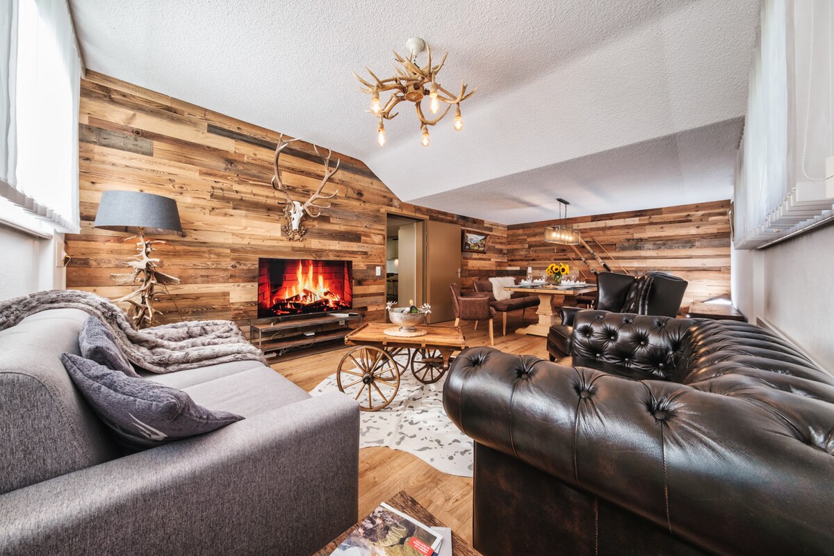 A cozy living space features a mix of rustic wood paneling and modern furnishings. A Chesterfield sofa complements a contemporary coffee table. A large TV displays a simulated fireplace, while antler decor adds character to the wall above. Large windows allow natural light to fill the room.