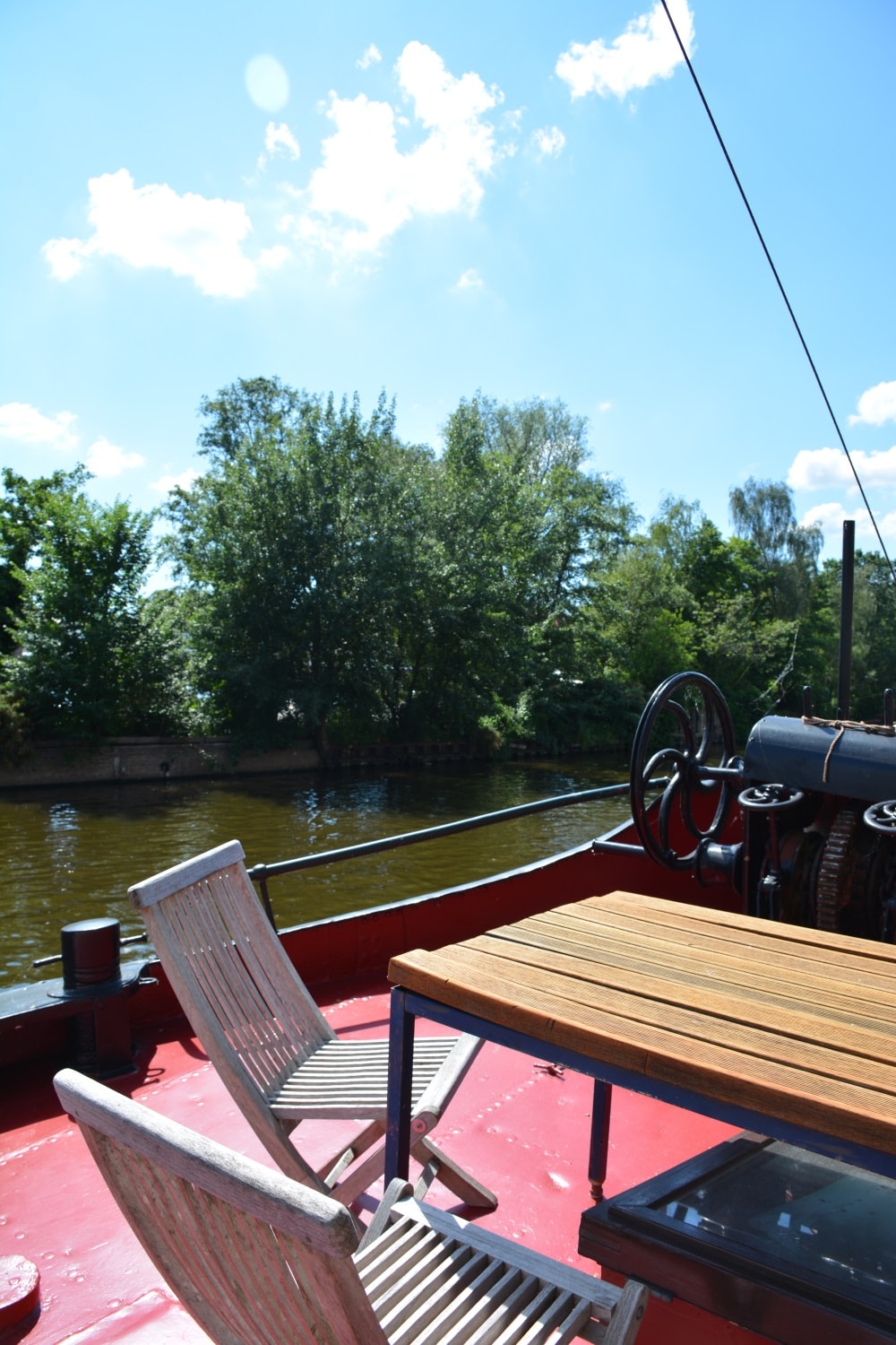 A serene outdoor space is presented on the boat's deck, featuring a wooden table accompanied by two white chairs. Lush greenery lines the banks of the canal, while the calm water reflects the blue sky dotted with soft clouds.