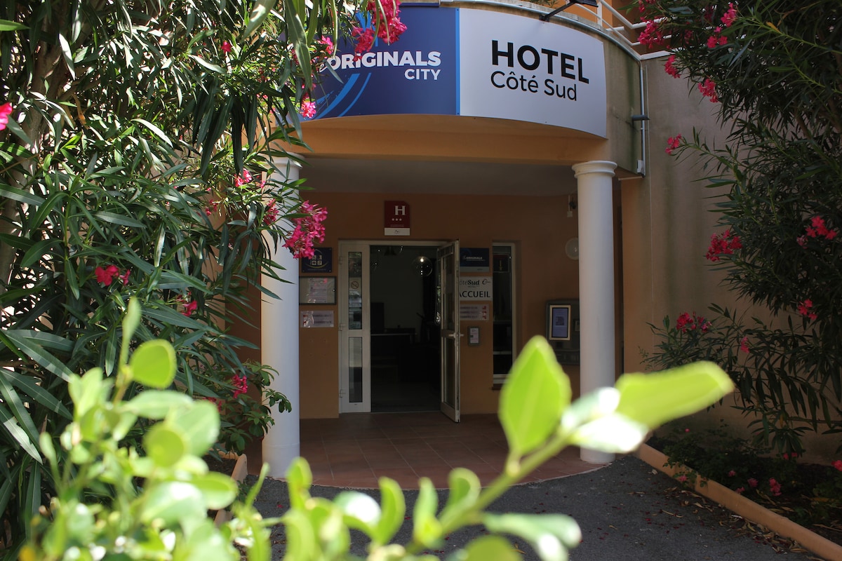 The hotel entrance is framed by lush greenery and colorful flowers, leading guests toward the inviting front door. Clear signage indicates 'HOTEL Côté Sud,' welcoming visitors into the establishment. A warm, approachable environment is presented, with spacious outdoor areas visible.