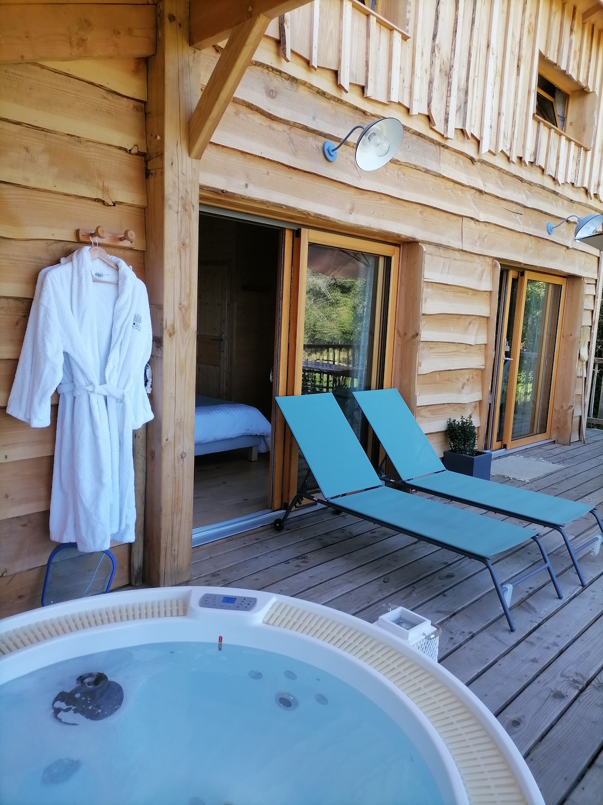 A private terrace features a hot tub, two lounge chairs, and a soft robe hanging on the wall. Large glass doors provide a view into the cozy interior, where a spacious bed is located. Wooden siding enhances the overall rustic charm.