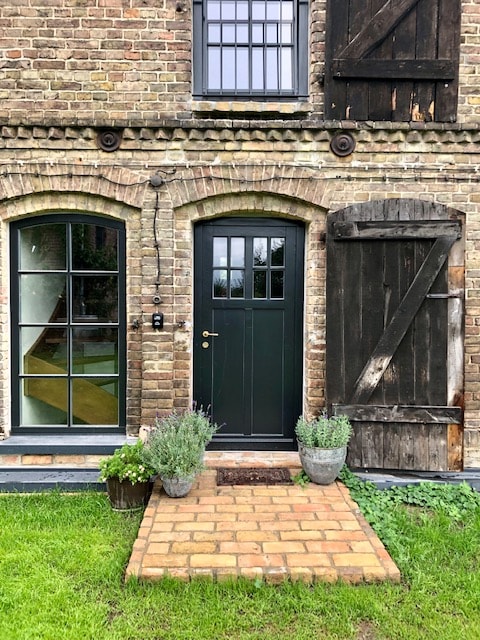 A charming entryway features a dark green door framed by two large windows with black divides. The rustic brick facade is complemented by a wooden barn door. A stone pathway leads to the door, flanked by potted plants, creating an inviting entrance.
