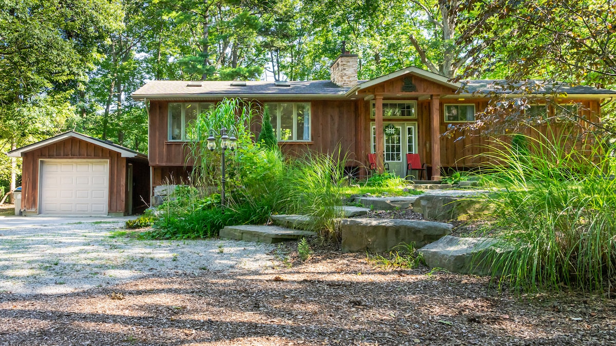 A charming vacation home is nestled among tall trees, featuring a stone pathway leading to the front entrance. The facade is characterized by warm wooden tones and large windows, allowing natural light to brighten the interior. A red chair set offers a welcoming spot for relaxation.