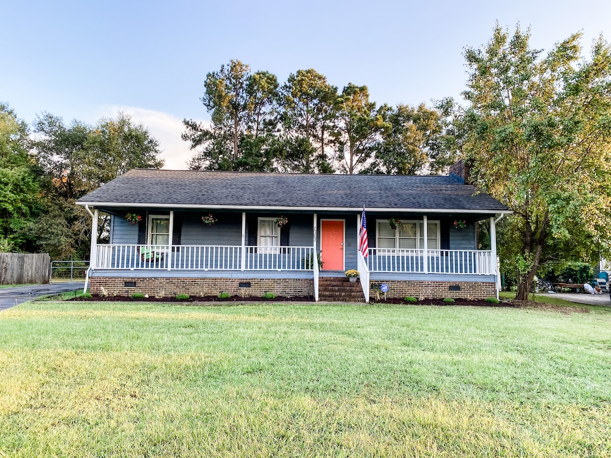 A single-story house is shown with a welcoming front porch adorned with flower baskets. An American flag is displayed beside the entrance. The lawn is well-maintained, bordered by a driveway. Tall trees provide a natural backdrop to the property.