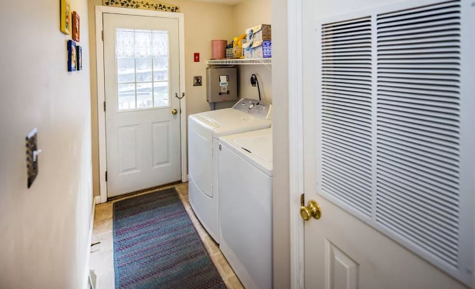 A laundry room is visible, featuring a washer and dryer positioned along the wall. A door provides access to the outside, while a textured rug adds comfort underfoot. Shelving above the appliances offers storage for laundry essentials.