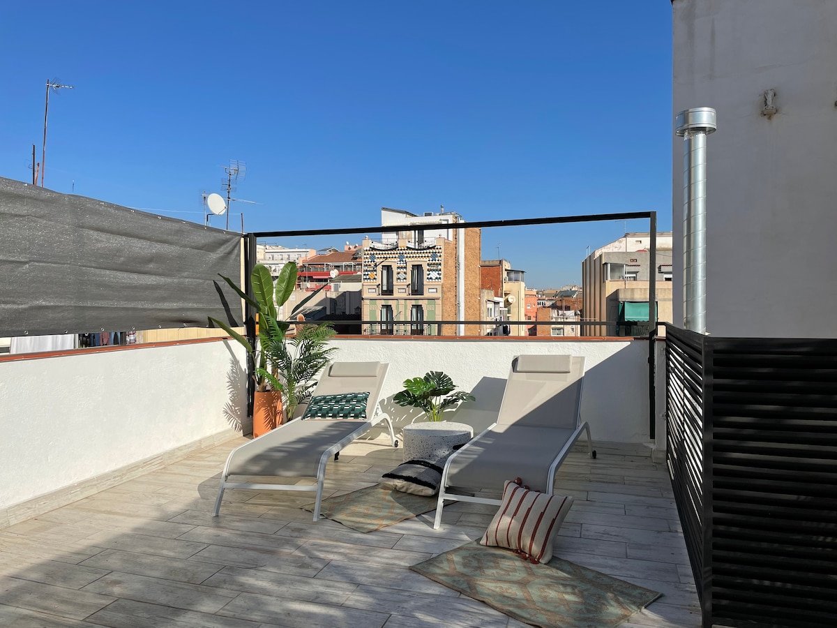 A spacious rooftop terrace is presented, featuring two loungers positioned under the clear blue sky. Decorative plants add a touch of greenery, while the surrounding cityscape is visible in the background. A privacy screen is partially seen, enhancing the relaxation area.