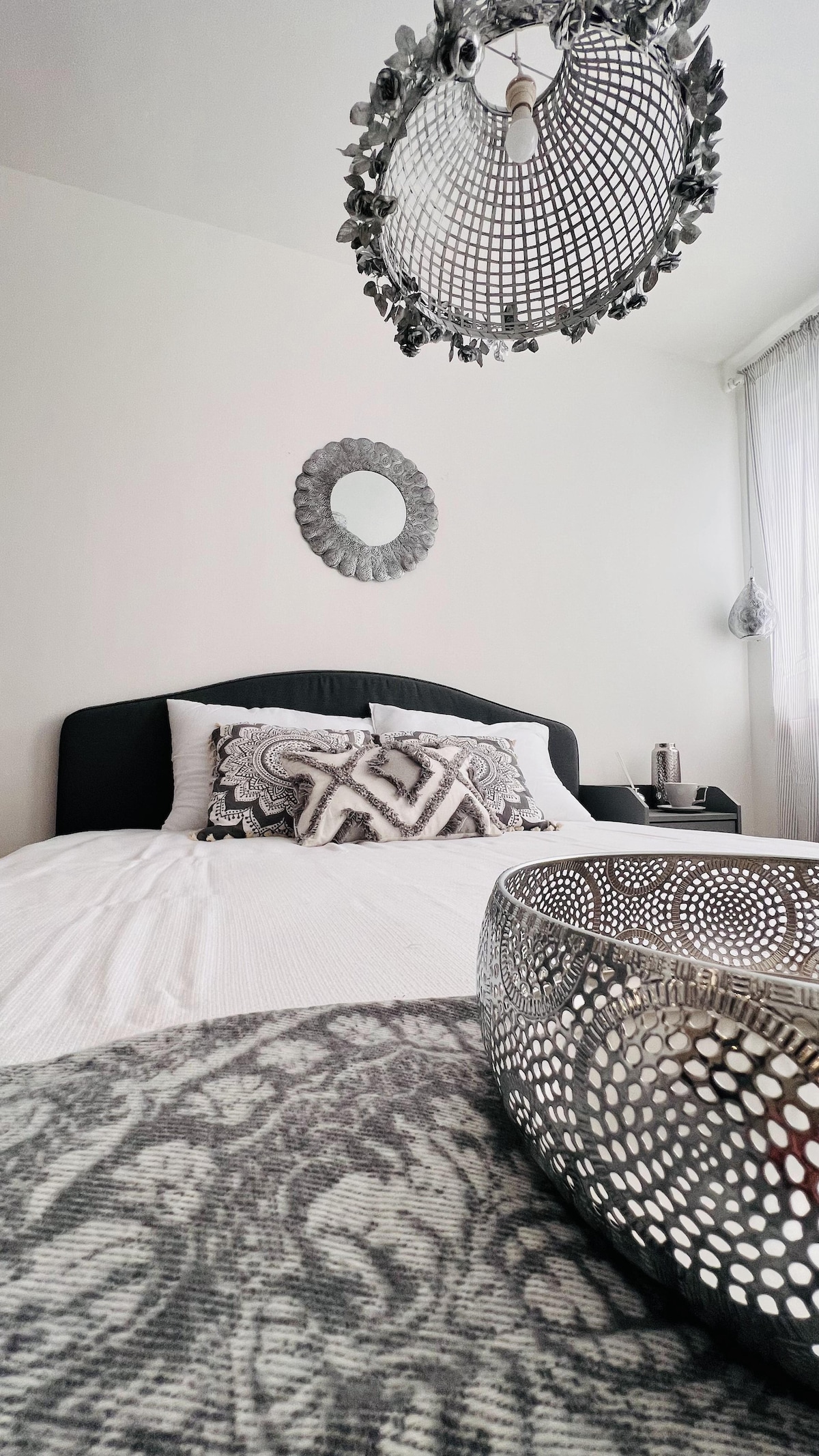 A spacious bedroom is presented with a large bed adorned with decorative pillows. A stylish light fixture hangs centrally from the ceiling, while a unique mirror complements the minimalist decor. A decorative bowl is placed on the bedspread, enhancing the elegant presentation.